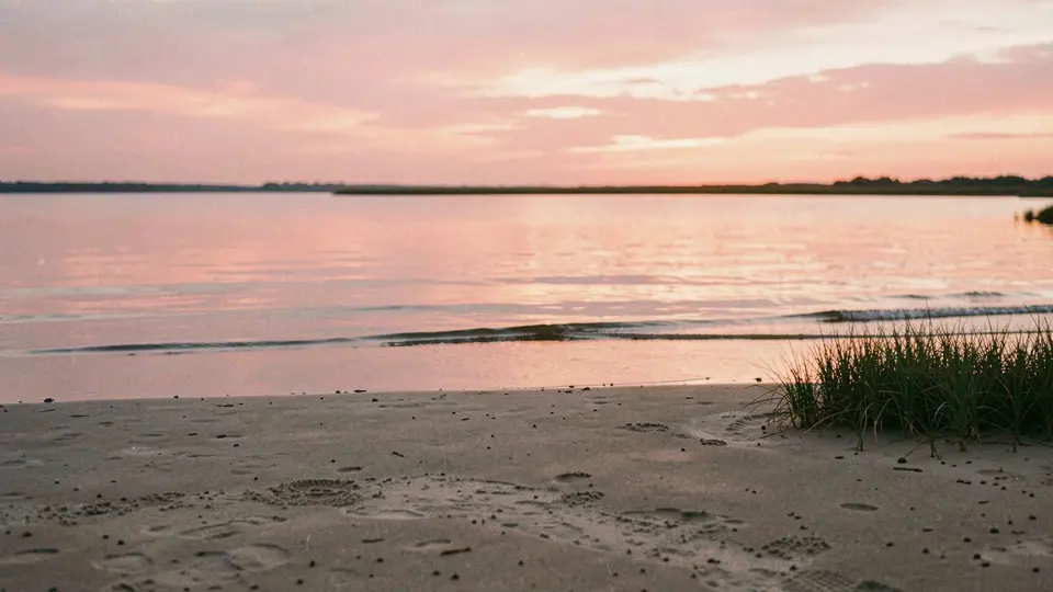 Napeague Bay at sunset, perfectly still water mirroring a pink and orange sky, a sand flat in the foreground, a low line of marsh grass.