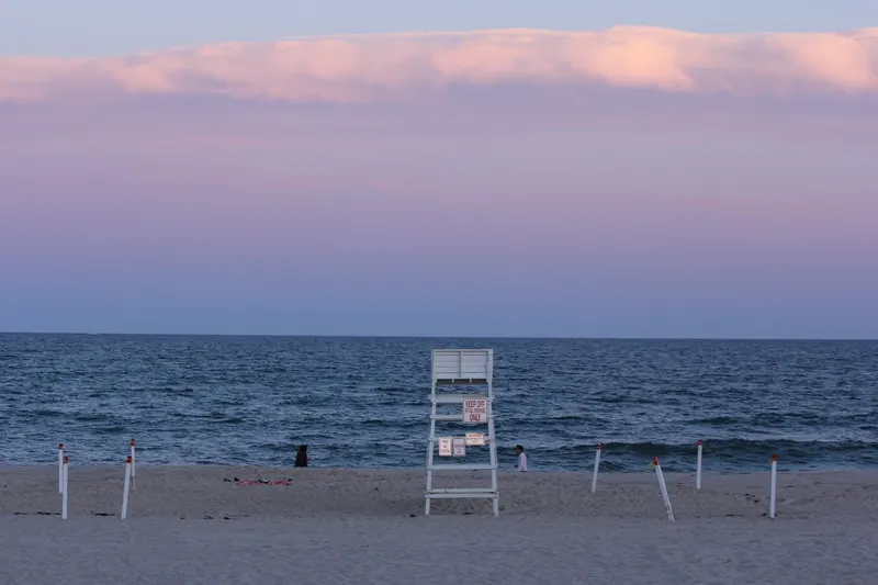 A lifeguard stand and roped stakes on an empty ocean beach at dusk under a pink sky.