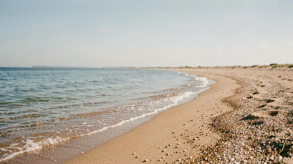 A long bay shoreline curving away, calm shallow water, mid-day sun, a sandy beach with fine pebbles, wide sky.