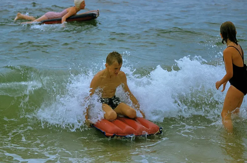 A boy kneels on a red inflatable raft in white surf while a girl and a woman in a swim cap stand behind in the water, in a faded color slide.