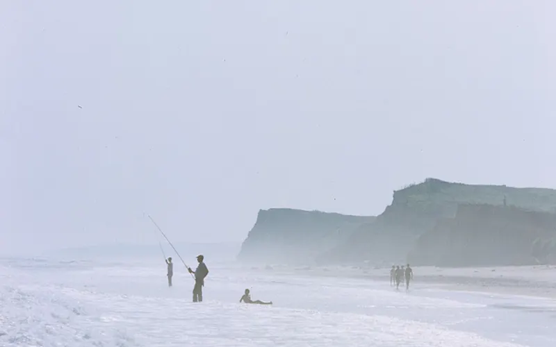 Two surf fishermen and a child stand in foam beneath tall hazy bluffs in a faded color slide.