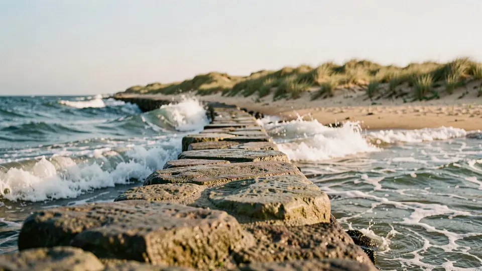 A stone groin jetty running out into rolling surf, dune grass behind, warm afternoon light and a pale blue sky.