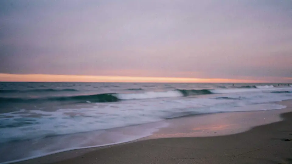 Blue hour on an empty beach, long-exposure waves smoothed into silk, lavender sky with a thin line of orange at the horizon.