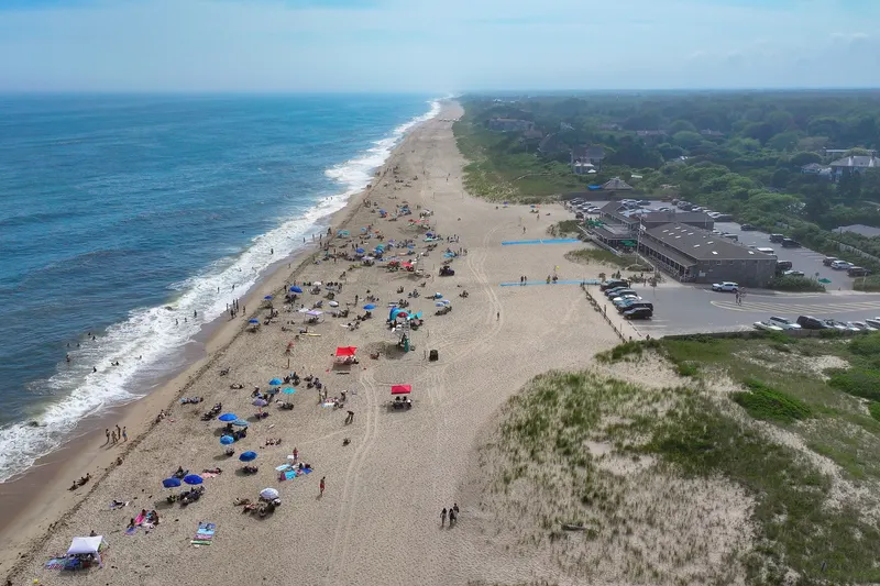 Aerial view of Main Beach in East Hampton with umbrellas on the sand, breaking surf, dune grass, the beach pavilion and a parking lot.