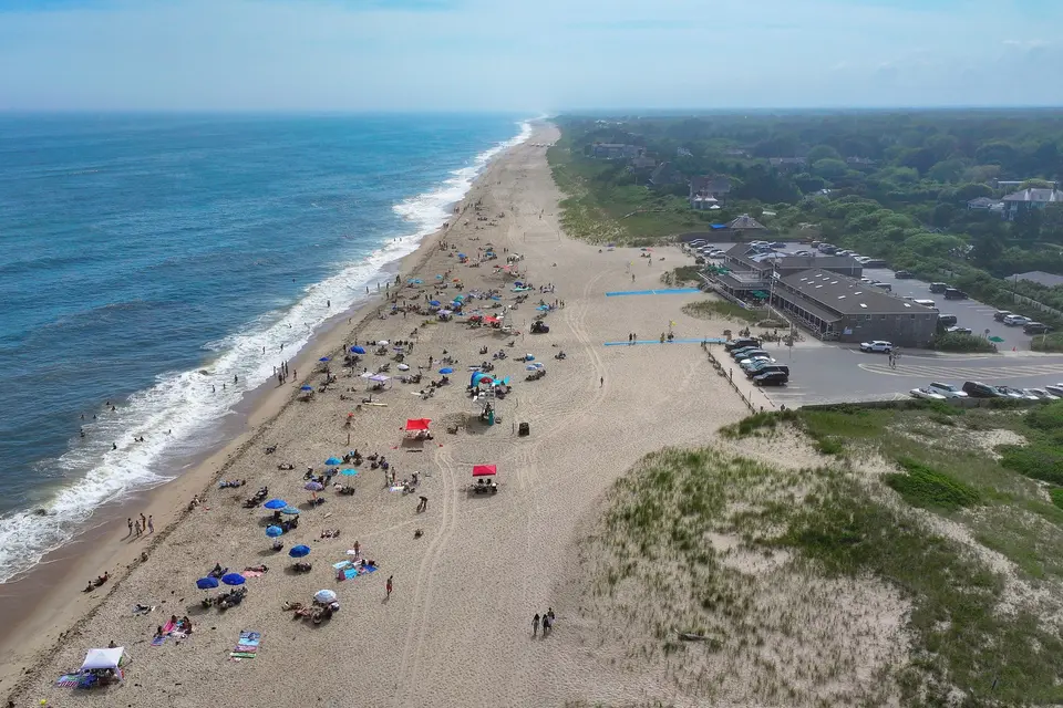 Aerial view of Main Beach in East Hampton with umbrellas on the sand, breaking surf, dune grass, the beach pavilion and a parking lot.