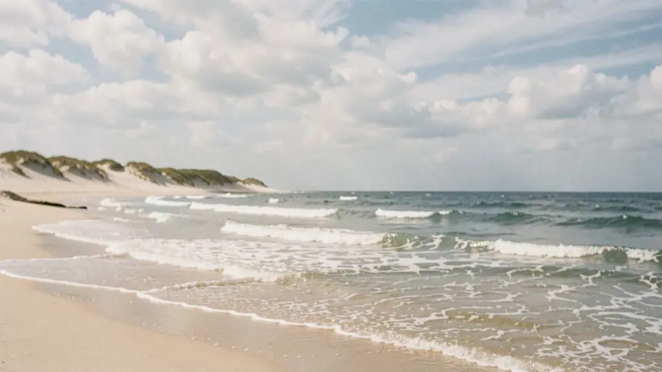 Pale beach and low dunes near an inlet at dusk, a salmon-pink sky over smooth silvery water, tiny waves, hushed.