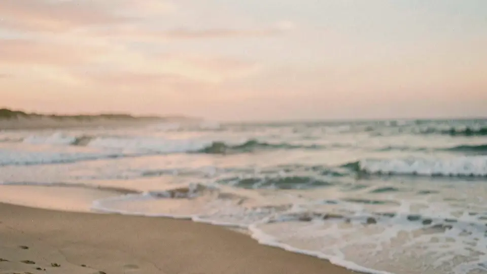 A soft pastel evening on a dune-backed ocean beach, long flat waves, creamy sky of peach and pale blue.
