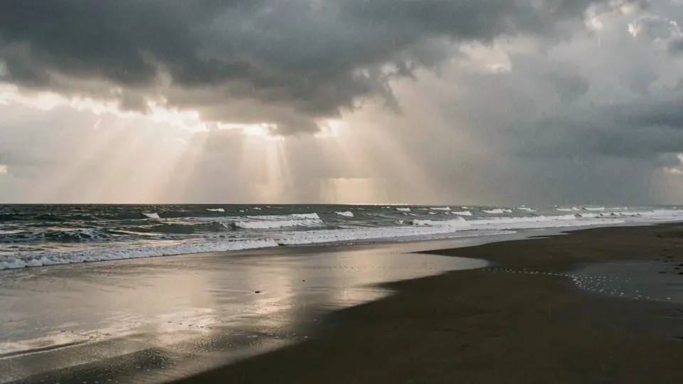 A storm clearing over the Atlantic, shafts of light through big clouds, a rain squall out at sea, wide dark wet beach.