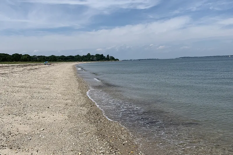 A curving pebble and sand bay beach with calm water, a wooded point and sailboats offshore.