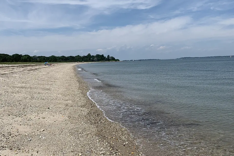 A curving pebble and sand bay beach with calm water, a wooded point and sailboats offshore.